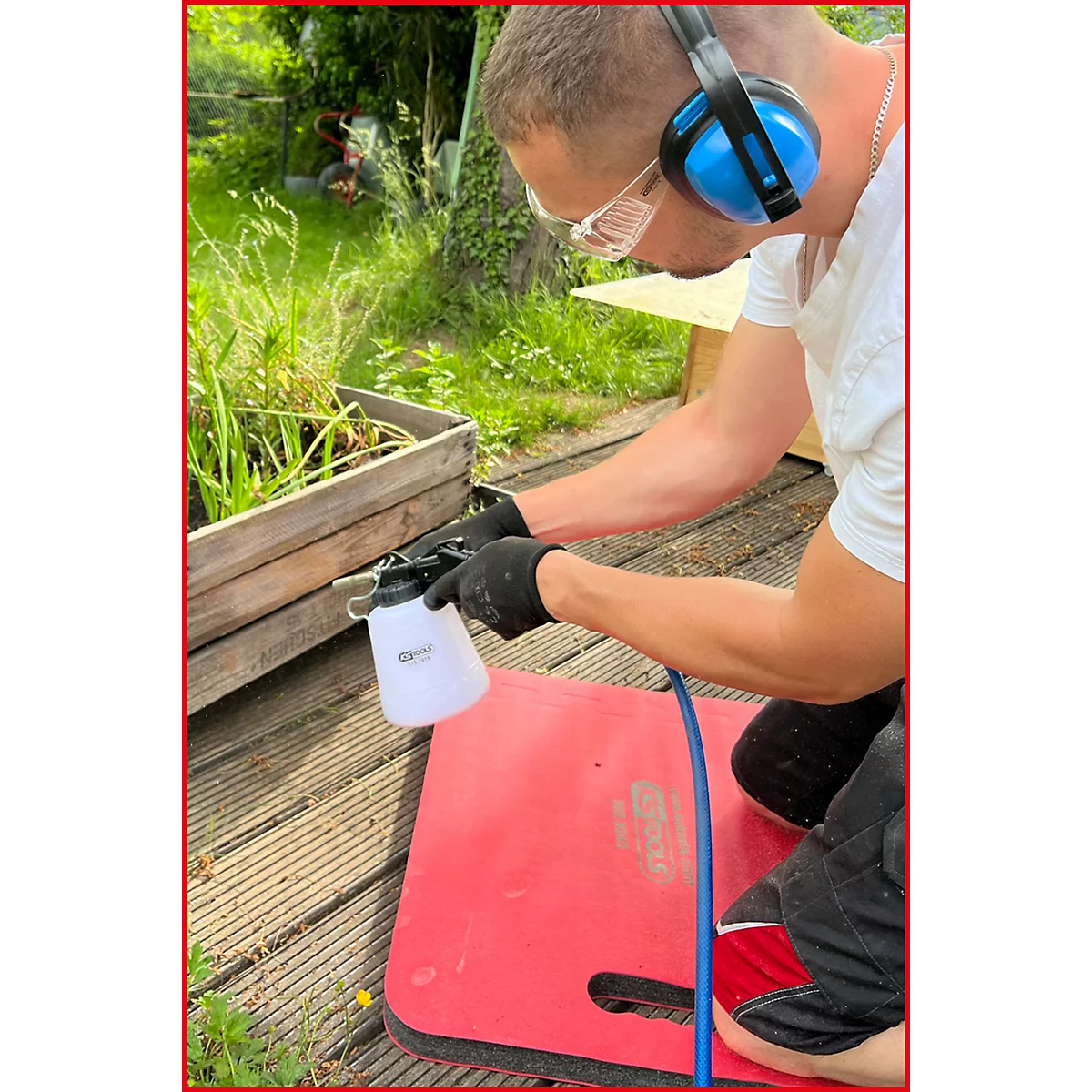 Ein Mann mit Gehörschutz und Brille kniet draußen und besprüht ein Holzkonstrukt mit einer Sprühflasche.